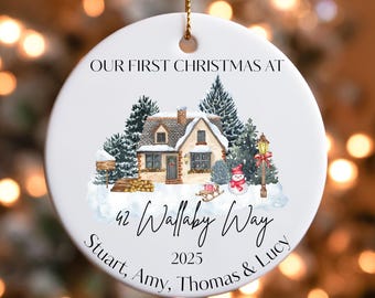 May include: White ceramic Christmas ornament with a winter scene featuring a house, trees, and a snowman. Text reads "OUR FIRST CHRISTMAS AT 42 Wallaby Way 2025" and names "Stuart, Amy, Thomas & Lucy". Includes a gold string for hanging.