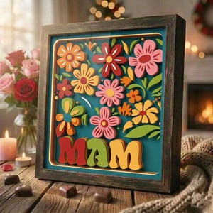 May include: A framed floral art piece with a teal background and colorful layered paper flowers. The word "MAM" is displayed in bold, layered letters. The frame is dark wood, and the artwork is on a wooden surface.