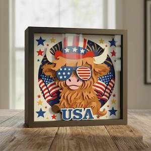 May include: A framed 3D paper art piece featuring a Highland cow wearing sunglasses with the American flag design. The cow is in front of a blue circle with American flags and stars. The word "USA" is at the bottom.