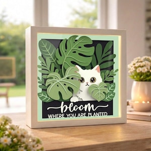 May include: A white framed art piece featuring a white cat peeking through green paper cut-out leaves. The art piece has the words "bloom where you are planted" in a cursive font on a black background. The frame is white.