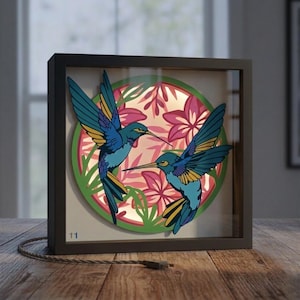 May include: A framed light box featuring two blue hummingbirds with yellow accents, set against a backdrop of pink flowers and green foliage. The box has a black frame and a USB cable. The artwork is illuminated from within.