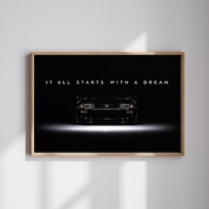 May include: A framed horizontal wall art print featuring a black Toyota Supra sports car centred against a dark background. The text IT ALL STARTS WITH A DREAM is printed in white capital letters above the vehicle, illuminated by a bright light.