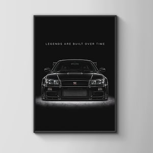 Puede incluir: Un póster con marco negro que muestra un coche deportivo Nissan GT-R negro de frente, centrado sobre un fondo oscuro. El texto "LEGENDS ARE BUILT OVER TIME" está impreso en letras mayúsculas blancas sobre el vehículo. Los faros son visibles.