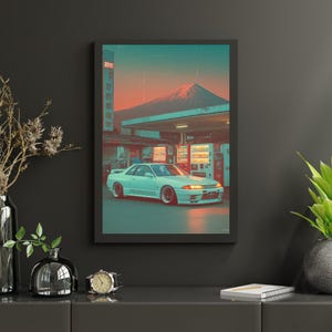 May include: A framed print featuring a light blue sports car parked in front of a Japanese gas station with vending machines. The background shows a mountain under a colorful sunset. The print is in a black frame.