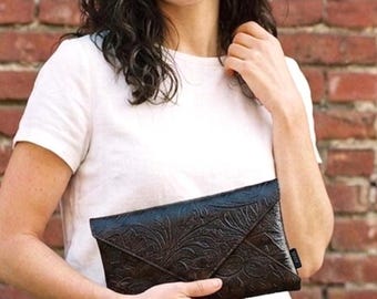 Op de afbeelding: Een zwarte clutch met een reliëf bloemenontwerp wordt vastgehouden door een persoon die een wit t-shirt en een blauwe spijkerbroek draagt. De achtergrond is een rode bakstenen muur. De clutch lijkt van leer te zijn.