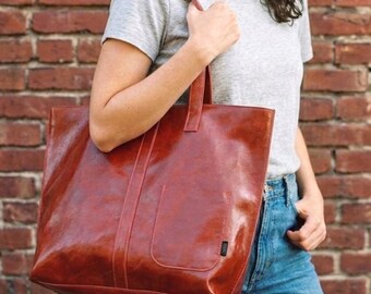 Op de afbeelding: Een grote, roodbruine leren tote bag met een rechthoekige vorm en een voorvak. De tas heeft twee handvatten en wordt vastgehouden door een persoon die een grijs t-shirt en een blauwe spijkerbroek draagt. De achtergrond is een bakstenen muur.