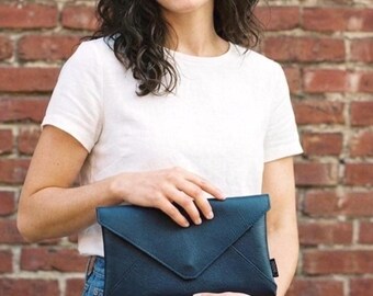 Op de afbeelding: Een vrouw houdt een donkerblauwe envelop-clutch vast. De clutch is gemaakt van een glad materiaal en heeft een eenvoudig, minimalistisch ontwerp. De vrouw draagt een wit t-shirt en een blauwe jeans, staand voor een rode bakstenen muur.