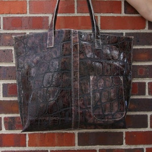 May include: Brown tote bag with a crocodile skin texture. The bag has a rectangular shape with a front pocket and two handles. The stitching is white. The bag is held in front of a brick wall.