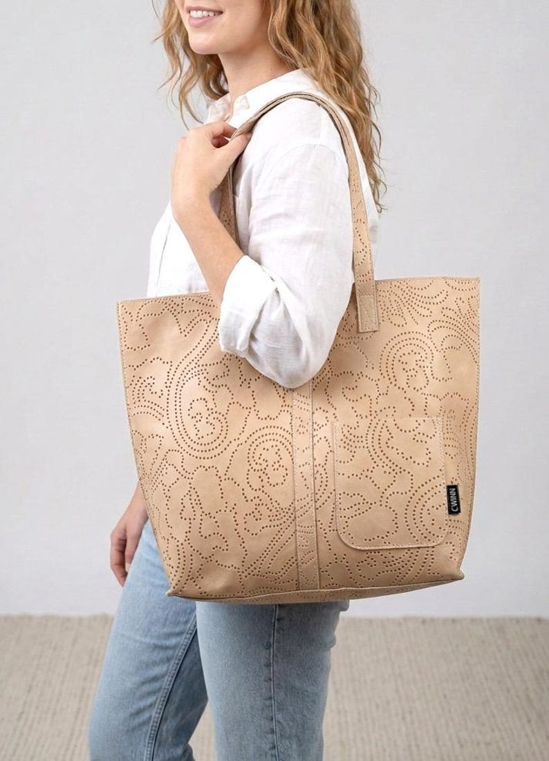 May include: Beige tote bag with a laser-cut floral design. The bag has a rectangular shape, a small pocket on the front, and two handles. The bag is made of a light-colored material.