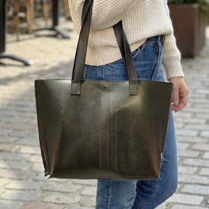 May include: Olive green tote bag with a rectangular shape and a front pocket. The bag has two handles and is being carried by a person wearing a cream-colored sweater and blue jeans. The person is wearing brown boots.