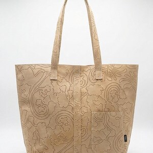 May include: A beige tote bag with a floral pattern cut into the leather. The bag has two handles and a front pocket. The bag is made of a light-colored leather.