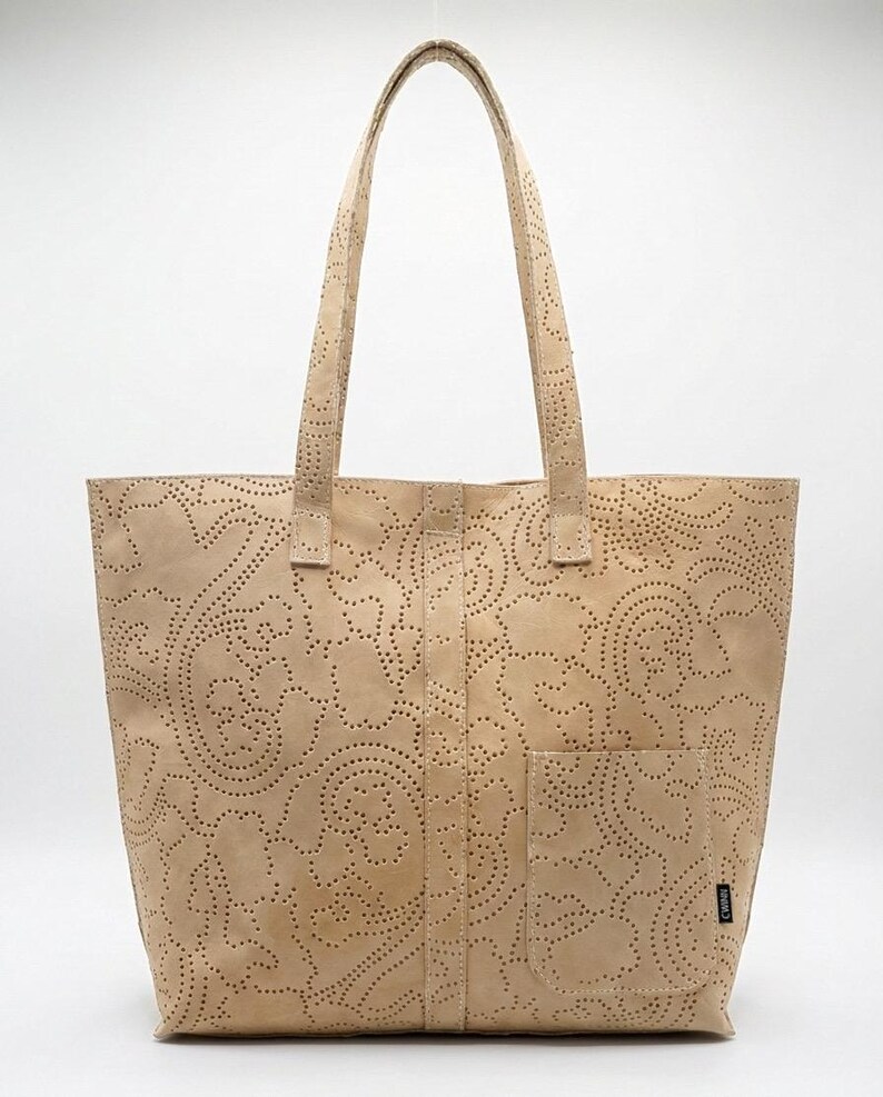 May include: A beige tote bag with a floral pattern cut into the leather. The bag has two handles and a front pocket. The bag is made of a light-colored leather.