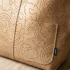 May include: A tan leather tote bag with a perforated paisley design. The bag has a small pocket on the front and a handle. The bag is made of a soft, supple leather and has a classic, timeless look.