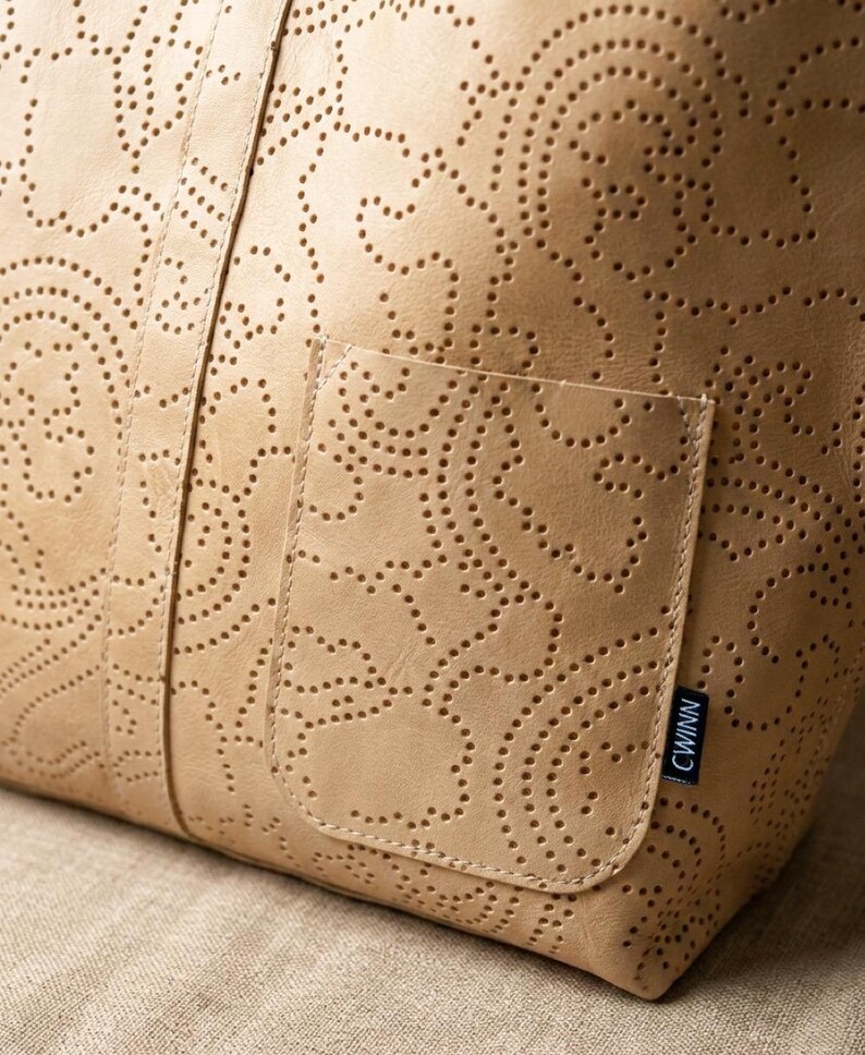 May include: A tan leather tote bag with a perforated paisley design. The bag has a small pocket on the front and a handle. The bag is made of a soft, supple leather and has a classic, timeless look.