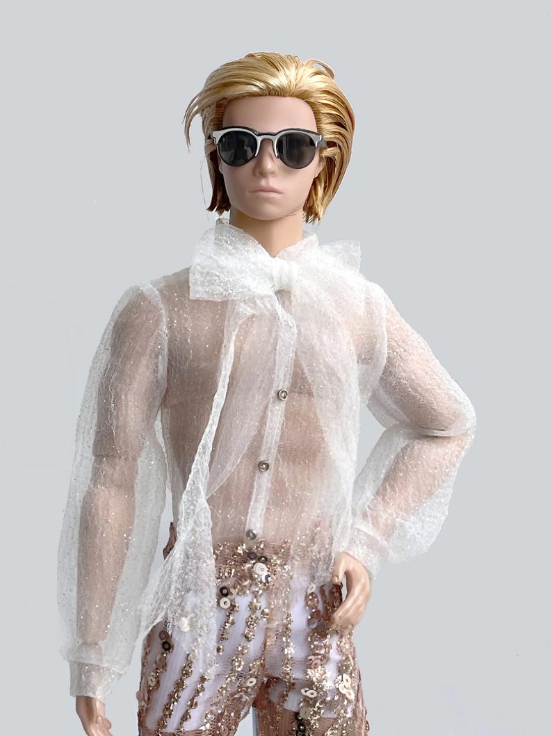 May include: A doll wearing a sheer white button-down shirt with a large bow and sequined pants. The doll has blonde hair and is wearing sunglasses. The pants are white with gold sequins. The shirt has long sleeves.