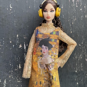 Peut inclure: Une poupée vêtue d'une robe dorée ornée d'une peinture de Gustav Klimt et d'un casque jaune. La poupée a les cheveux bruns et est posée devant un fond sombre et texturé. Une machine à écrire vintage est visible au premier plan.