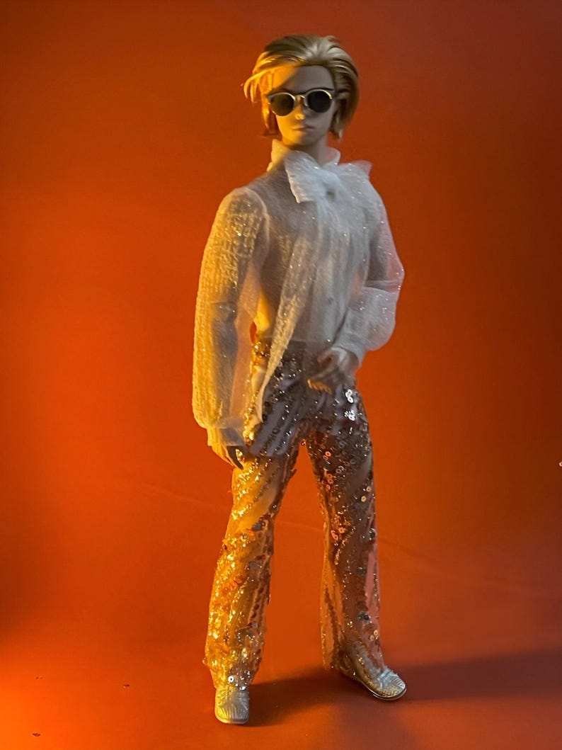 May include: A doll wearing a sheer white shirt, sequined pants, and sunglasses. The doll has blonde hair and is posed against an orange background. The pants are gold with silver sequins.