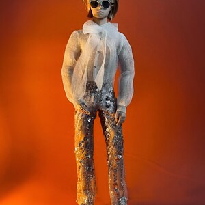 May include: A doll wearing a sheer white blouse with a large bow, sunglasses, and sequined pants. The doll has short blonde hair and silver sneakers. The background is a gradient of orange and red.