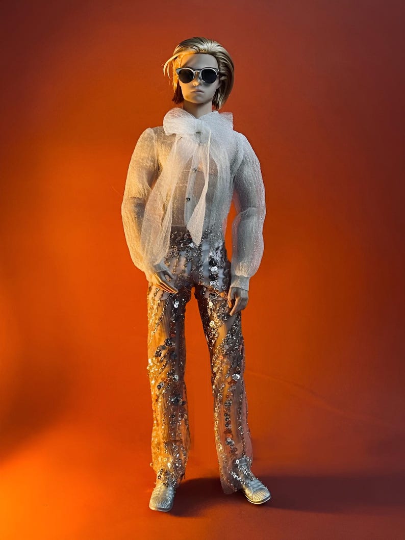 May include: A doll wearing a sheer white blouse with a large bow, sunglasses, and sequined pants. The doll has short blonde hair and silver sneakers. The background is a gradient of orange and red.