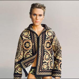 Puede incluir: Una muñeca con una camisa abotonada con estampado negro y dorado y ropa interior a juego. La camisa presenta un diseño repetido de medallones y volutas. La muñeca tiene cabello rubio y ojos azules.
