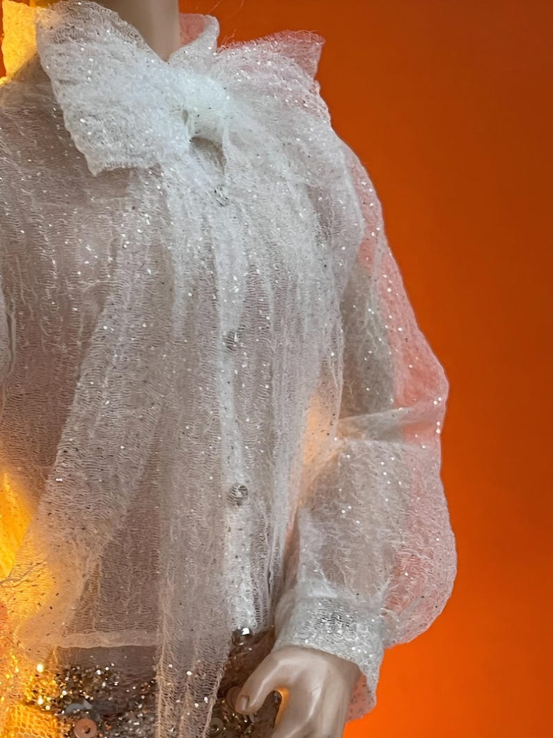 May include: A white, sheer, long-sleeved blouse with a large, ruffled collar and cuffs. The blouse is embellished with sequins, adding a touch of sparkle. The buttons are also embellished. The garment is displayed against an orange background.