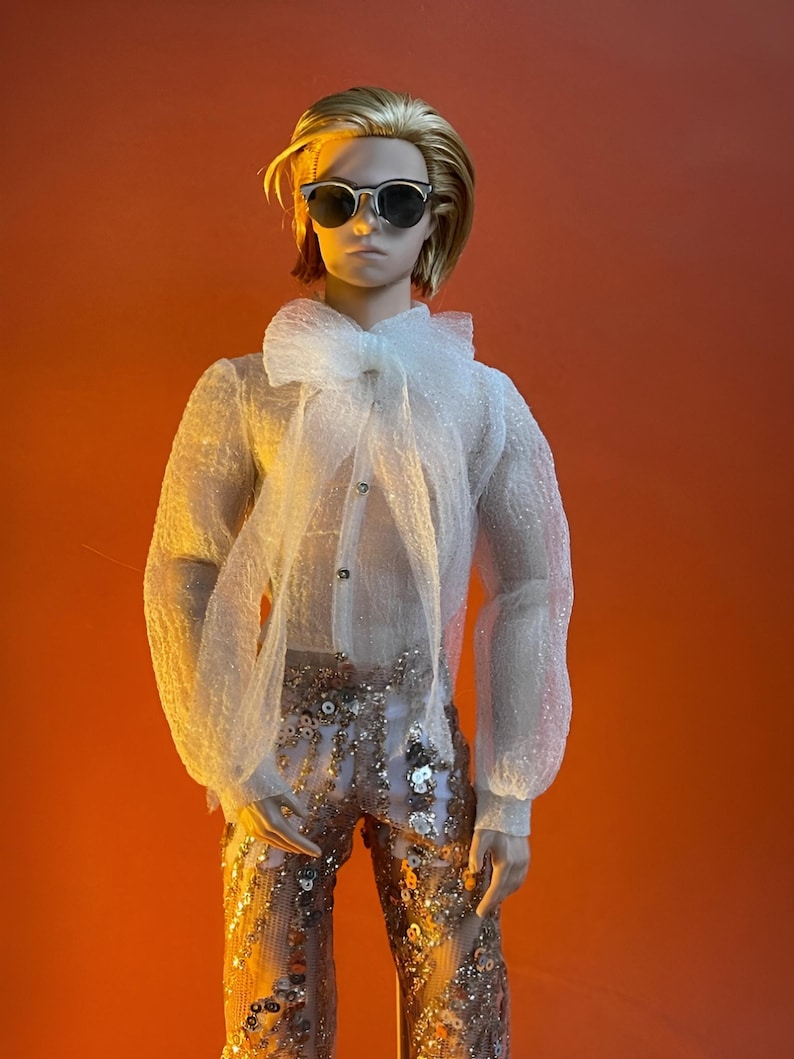 May include: A doll wearing a sheer white blouse with a large bow and sequined pants. The doll has sunglasses and blonde hair. The background is a solid orange color.