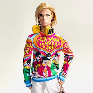 Könnte beinhalten: Eine Puppe mit blondem Haar trägt ein langärmeliges Sweatshirt mit einem bunten psychedelischen Muster, dem Text "ALL YOU NEED IS LOVE" und Illustrationen von vier Figuren. Die Puppe trägt gelbe Kopfhörer um den Hals und eine weiße Hose.