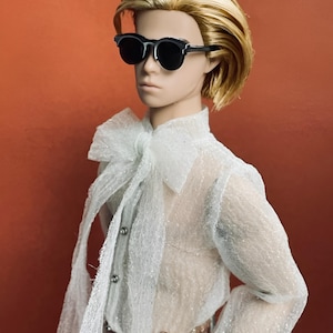 May include: A doll with short, blonde hair wearing black sunglasses, a sheer white shirt with a large bow, and a decorative belt. The doll is set against a solid orange background.