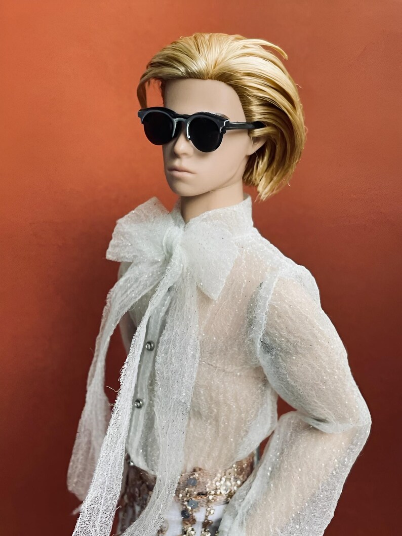 May include: A doll with short, blonde hair wearing black sunglasses, a sheer white shirt with a large bow, and a decorative belt. The doll is set against a solid orange background.