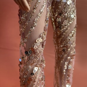 May include: Close-up of a doll's legs wearing a sheer, nude-colored jumpsuit. The jumpsuit is embellished with gold sequins and a delicate net-like fabric. The sequins are arranged in diagonal lines, creating a shimmering effect. The background is a solid peach color.