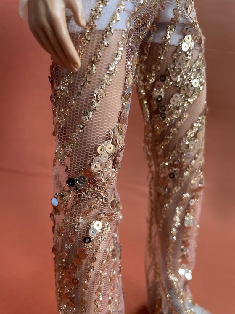 May include: Close-up of a doll's legs wearing a sheer, nude-colored jumpsuit. The jumpsuit is embellished with gold sequins and a delicate net-like fabric. The sequins are arranged in diagonal lines, creating a shimmering effect. The background is a solid peach color.