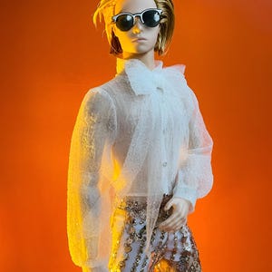 May include: A doll wearing a white, sheer, long-sleeved shirt with a ruffled collar and cuffs. The doll is also wearing gold sequined pants and black-rimmed sunglasses. The doll has blonde hair and is set against an orange background.