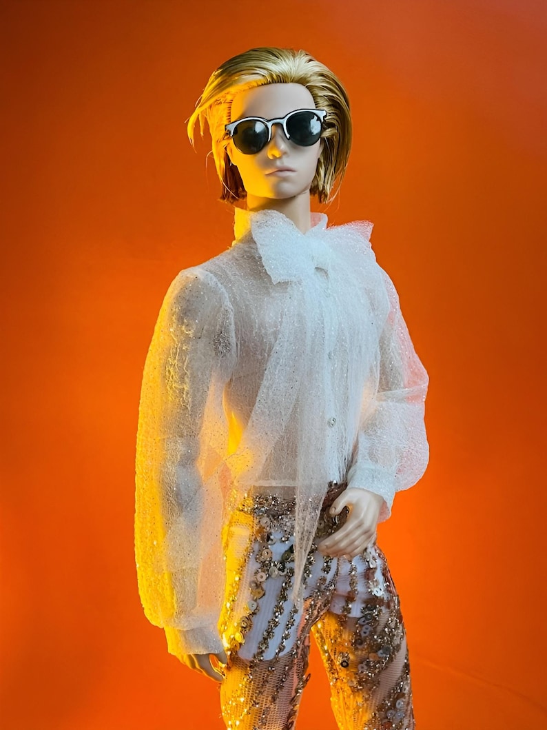 May include: A doll wearing a white, sheer, long-sleeved shirt with a ruffled collar and cuffs. The doll is also wearing gold sequined pants and black-rimmed sunglasses. The doll has blonde hair and is set against an orange background.
