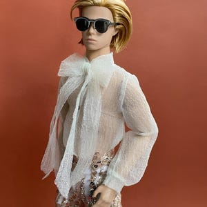 May include: A doll with blonde hair and sunglasses, wearing a sheer white blouse with a large bow and sequined pants. The doll is posed against a solid orange background.