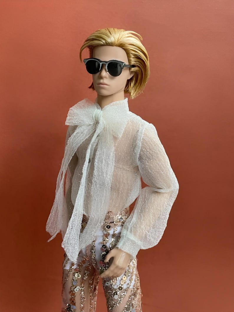 May include: A doll with blonde hair and sunglasses, wearing a sheer white blouse with a large bow and sequined pants. The doll is posed against a solid orange background.