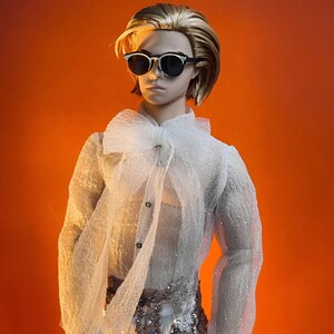 May include: A doll wearing sunglasses, a white sheer blouse with a large bow, and sequined pants. The doll has blonde hair and is set against an orange background. The blouse has long sleeves and button details.