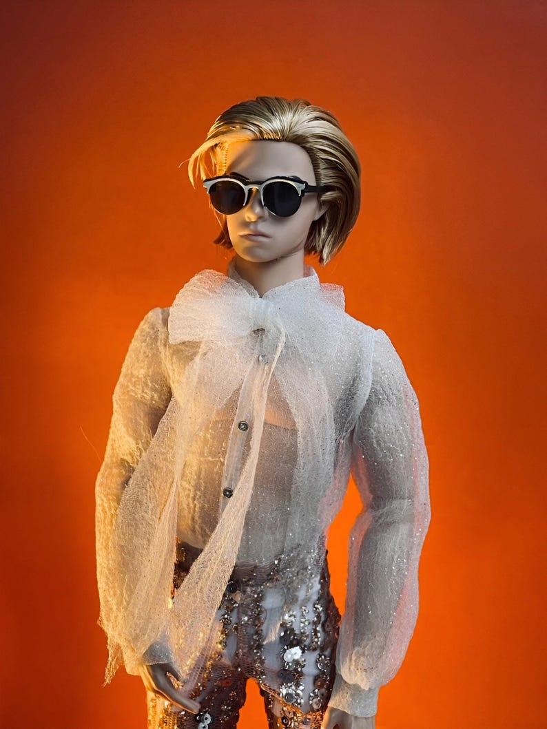 May include: A doll wearing sunglasses, a white sheer blouse with a large bow, and sequined pants. The doll has blonde hair and is set against an orange background. The blouse has long sleeves and button details.