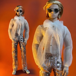 May include: A doll wearing a sheer white blouse with a large bow, sunglasses, and sequined pants. The doll has blonde hair and silver shoes. The background is a solid orange color. The doll is posed in front of a matching doll.