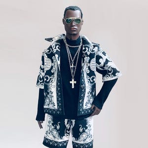 Könnte beinhalten: Eine Schaufensterpuppe trägt eine kurzärmelige Jacke mit schwarz-weißem Muster, einen schwarzen Rollkragenpullover und passende Shorts. Das Outfit wird durch eine silberne Kreuzkette und eine Sonnenbrille mit Goldrand ergänzt. Jacke und Shorts haben ein verziertes Scroll-Design.