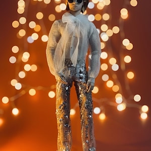 May include: A doll wearing a sheer white blouse with a large bow, sequined pants, and sunglasses. The doll is posed against a warm orange background with blurred string lights. The doll is wearing silver shoes.