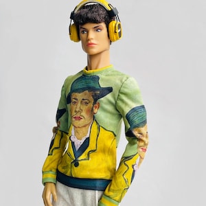 Puede incluir: Muñeca con auriculares amarillos y una camiseta verde de manga larga con una pintura de un hombre con una chaqueta amarilla y un sombrero verde. La muñeca también lleva pantalones blancos.