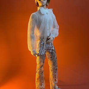 May include: A doll wearing a sheer white ruffled shirt, sequined pants, and silver shoes. The doll has short blonde hair and sunglasses. The background is a solid orange.