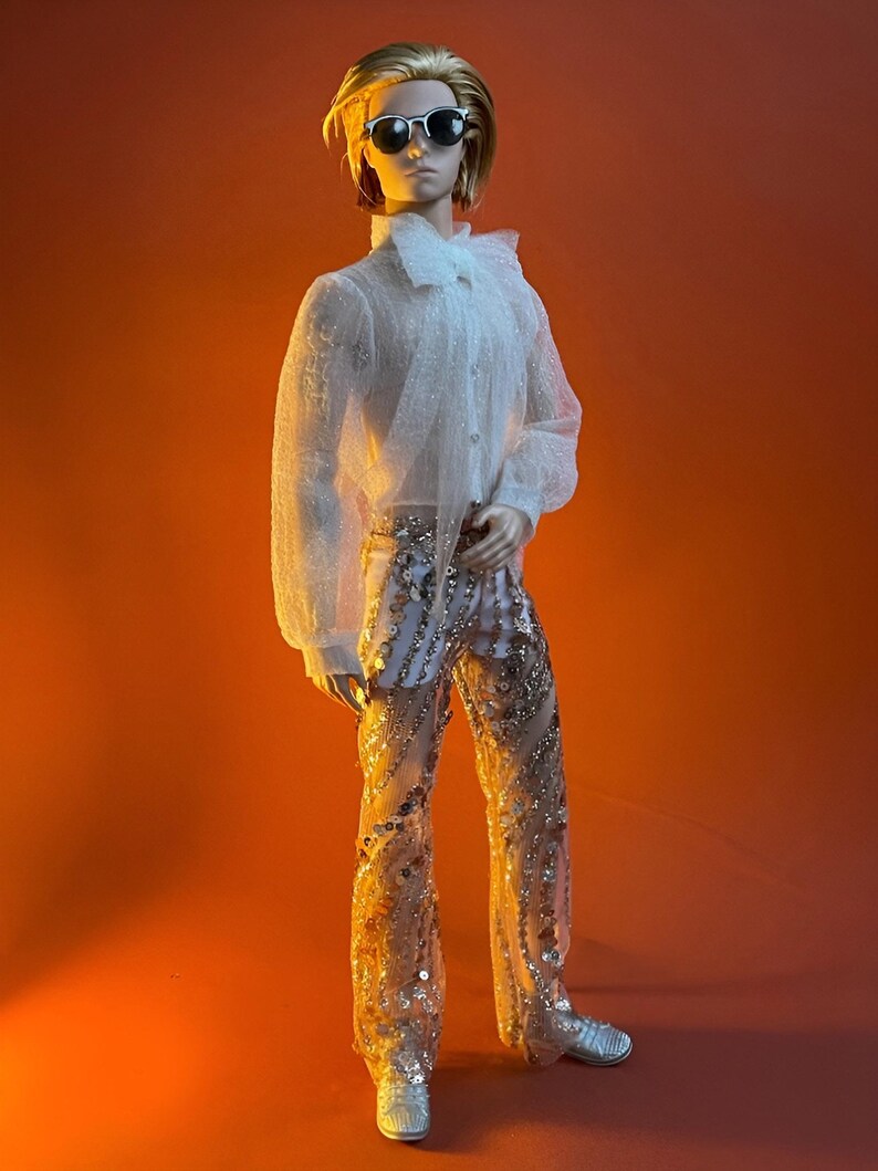 May include: A doll wearing a sheer white ruffled shirt, sequined pants, and silver shoes. The doll has short blonde hair and sunglasses. The background is a solid orange.