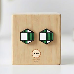 May include: A wooden block with two hexagonal green and white geometric designs, resembling eyes. Below the eyes are three small dots. The block is on a white surface, with a plant and a book in the background.