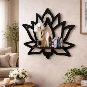 Può includere: Una mensola da parete nera a forma di fiore di loto con un ripiano marrone scuro. La mensola contiene un diffusore in vetro, cristalli e fiori decorativi. Il muro è di colore beige chiaro.
