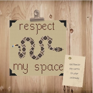 May include: Cross-stitch art with the words "respect my space" and a snake design. The snake is brown and tan, with a blue and yellow detail. The art is held by a silver clip on a wooden background.