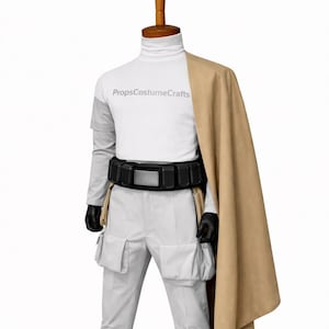 Puede incluir: Un disfraz en un maniqu&iacute;. Presenta una camiseta blanca de cuello alto con el texto "PropsCostumeCrafts", pantalones grises claros con bolsillos, un cintur&oacute;n negro, guantes negros, una capa color canela y zapatos negros. El disfraz se exhibe en un maniqu&iacute;.