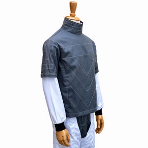 May include: A gray top with white stitching and a high collar, layered over a white long-sleeved shirt. The outfit includes white pants. The sleeves have black cuffs. The clothing is displayed on a wooden mannequin.