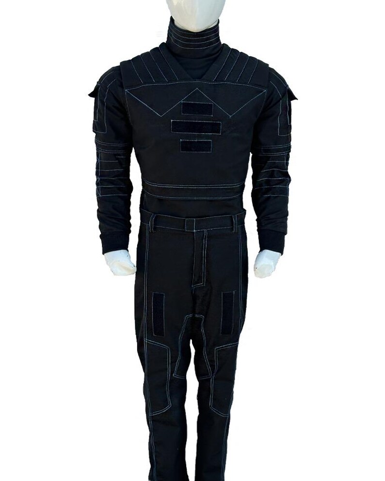 May include: Black jumpsuit with white stitching details. The outfit features a high collar, long sleeves, and multiple rectangular patches on the chest and legs. The jumpsuit is displayed on a mannequin.