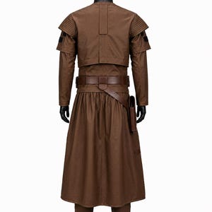 Puede incluir: Un disfraz marr&oacute;n con un abrigo largo, chaqueta y pantalones. La chaqueta tiene detalles en los hombros y un cintur&oacute;n con una funda. El disfraz se muestra en un maniqu&iacute; negro sobre un fondo blanco. El atuendo est&aacute; hecho de un material duradero.